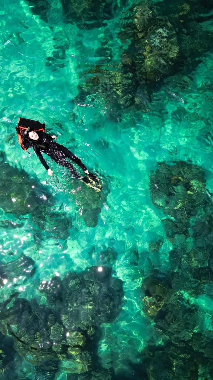 Eine Person schwimmt in klarem türkisblauem Wasser vor steinigen Felsen.