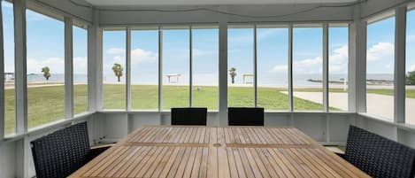 A covered patio with a wooden table and chairs, large windows offering a view of the beach.