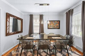 Ein Esszimmer mit einem rechteckigen Tisch, sechs Stühlen, einem großen Spiegel, einem farbenfrohen Gemälde und einem gemusterten Teppich.