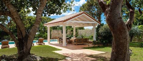 Un área de estar al aire libre con techo, piscina, árboles y una pared de piedra.