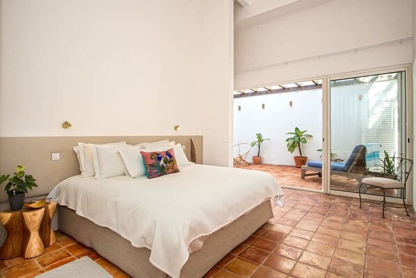 Un dormitorio con una cama grande, una puerta corrediza de vidrio que da a un patio con plantas en macetas y una silla.
