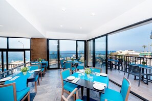 A restaurant with tables set for dining, large windows offering a view of the sea, and a balcony with outdoor seating.
