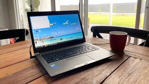 A laptop on a wooden table with a red mug beside it, a window with a view of a grassy area and a fence.