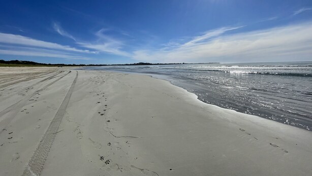 A sandy beach with footprints and tire tracks, leading to the ocean under a clear sky.
