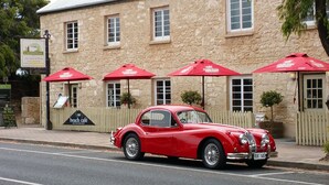 A vintage red car parked in front of a stone building with red umbrellas and a sign for 'Beach Café'.