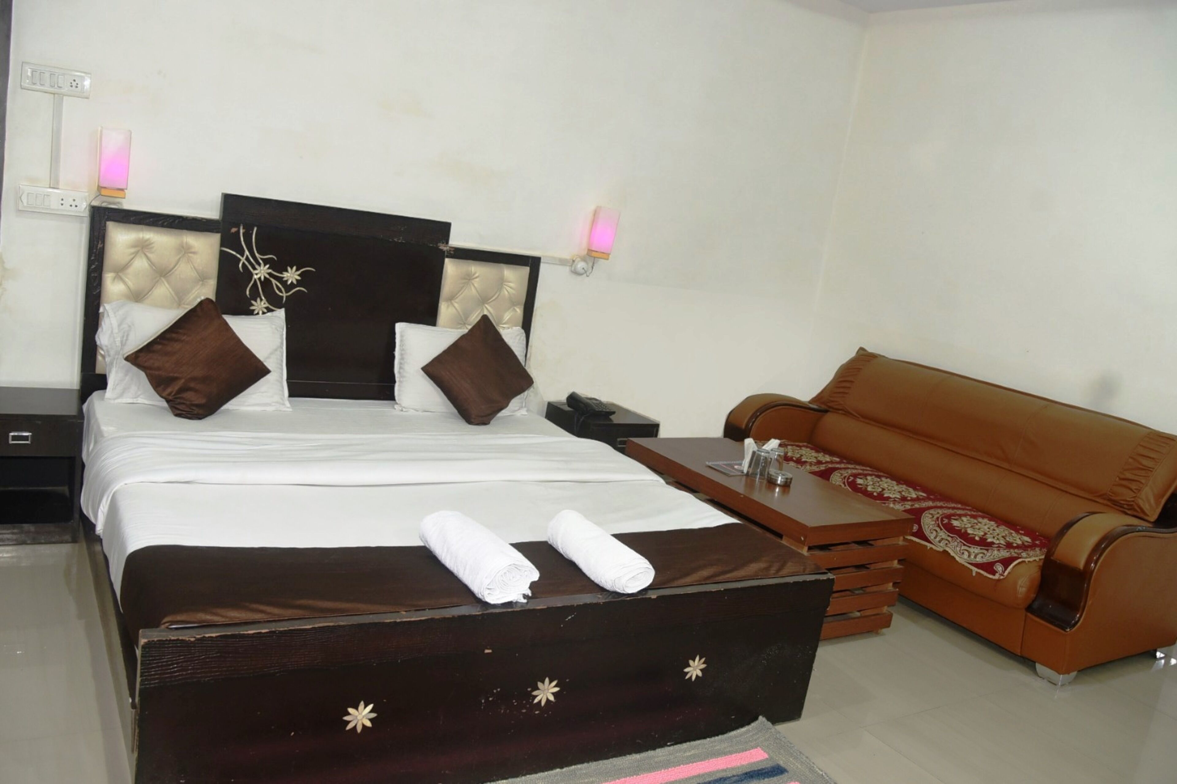 A hotel room with a bed, a wooden headboard, a brown sofa, a small table, and two bedside lamps.