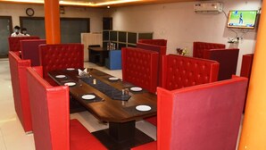 A restaurant with red upholstered booths, a long wooden table with plates and cutlery, and a TV mounted on the wall. - Hotel Udika Palace (Singrauli)
