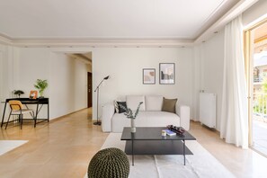 Un salon moderne avec un canapé blanc, une table basse noire et une petite table d’appoint. On y trouve un bureau avec une chaise et une lampe, ainsi qu’une plante posée sur le bureau. La pièce est dotée d’une grande fenêtre avec des rideaux.
