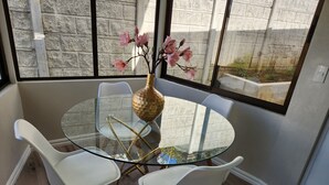 A round glass table with two white chairs, a vase with pink flowers, and a view of a stone wall through large windows. - Beautiful and cozy home in a safe and private environment (Alajuela)