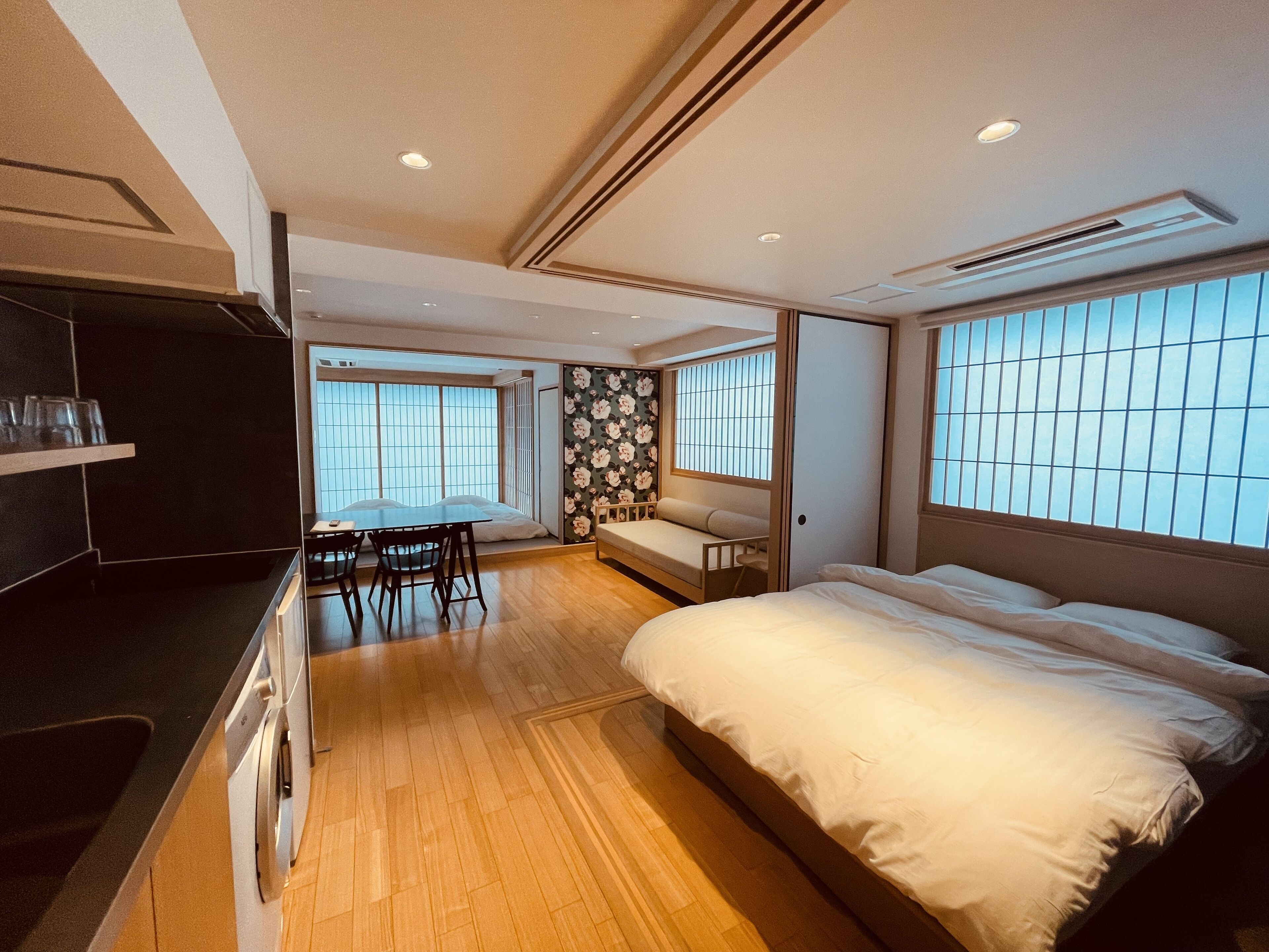 A modern hotel room with a wooden floor, a bed, a small table with chairs, and large windows with blinds.