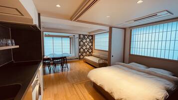 A modern hotel room with a wooden floor, a bed, a small table with chairs, and large windows with blinds.