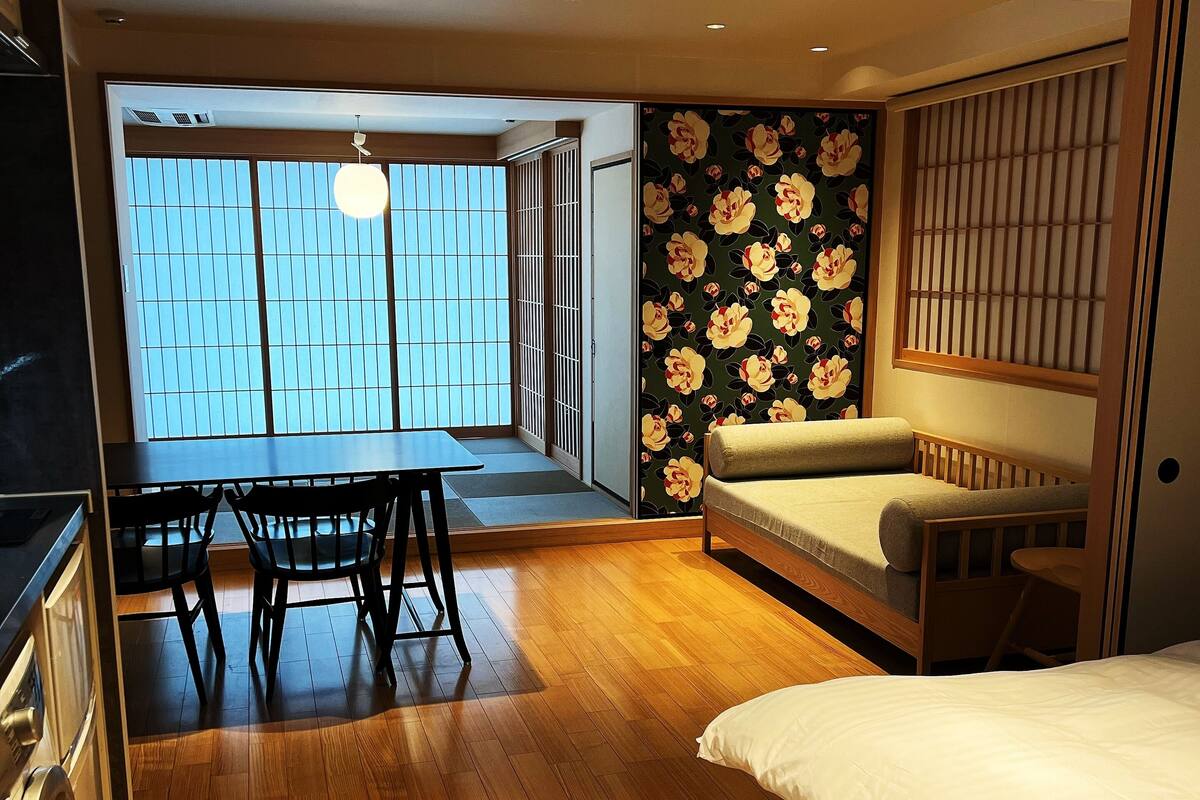 Una habitación de hotel moderna con suelo de madera, una cama, una mesa de comedor con sillas y vistas por una puerta corredera.