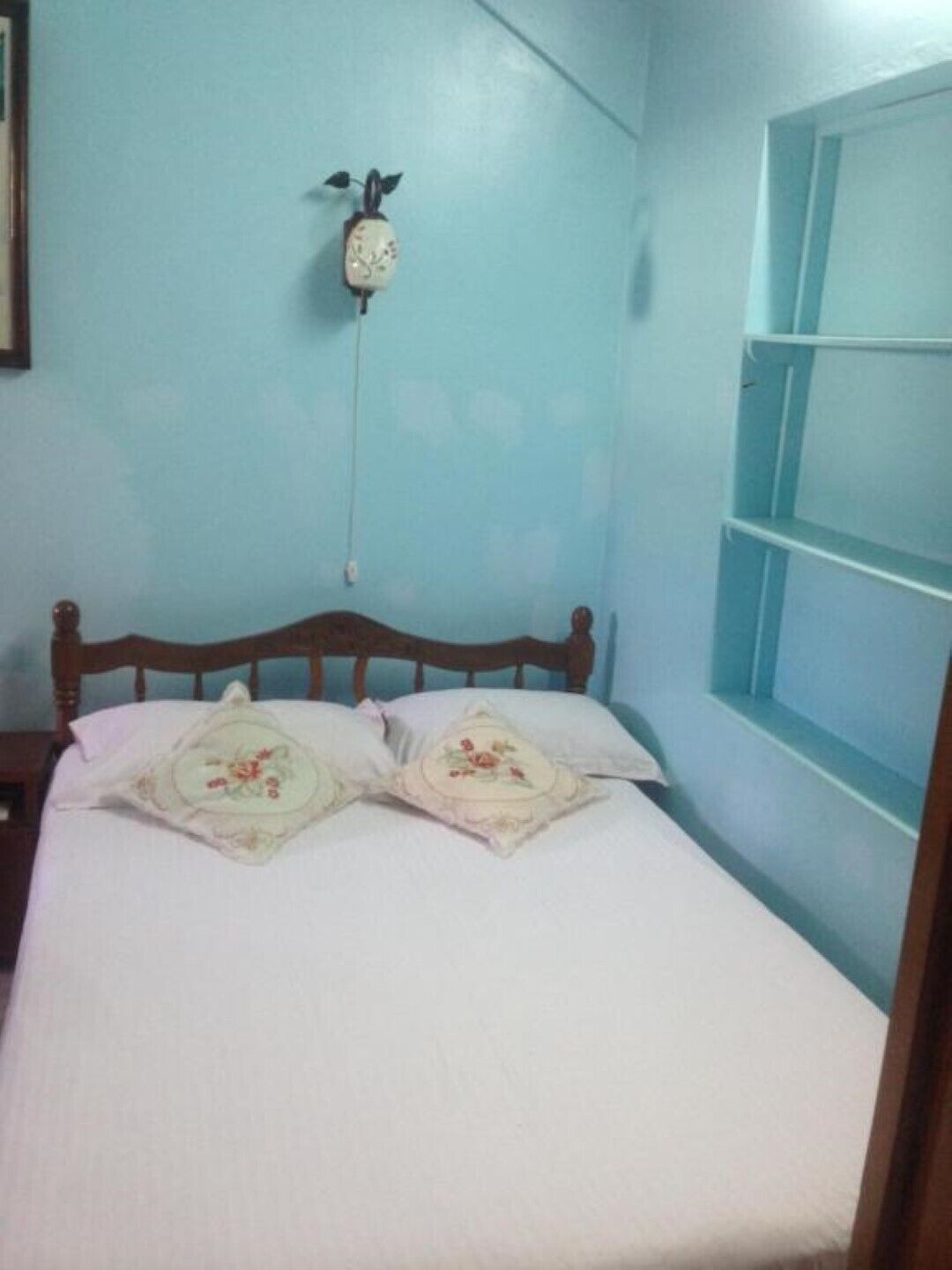 A bedroom with a wooden bed, floral pillows, a wall clock, and a window with a blue frame.