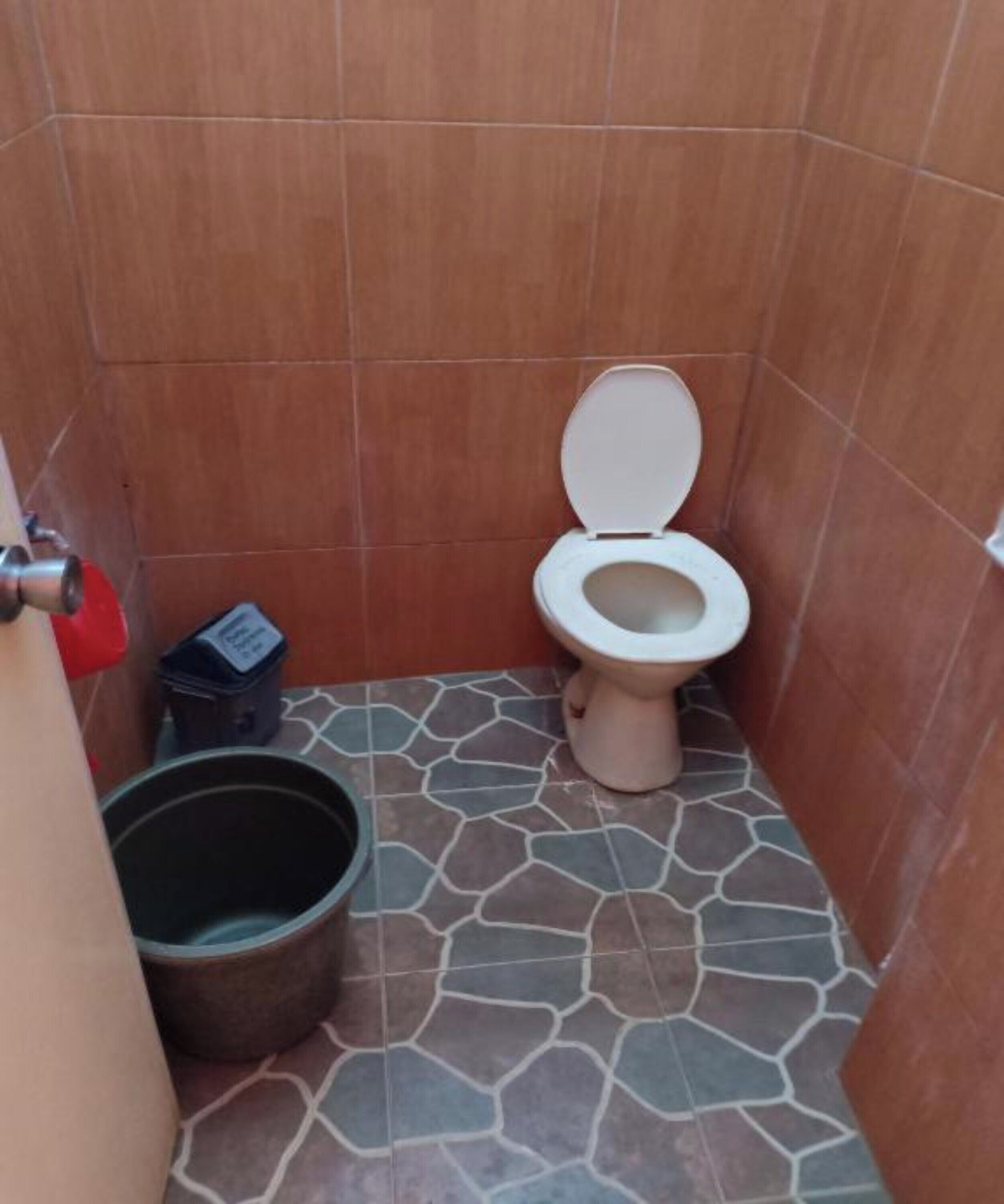 A small bathroom with a toilet, a red bucket, and a black trash bin.