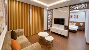 A modern hotel room with a sofa, two round tables, a TV, a bed, and a sliding door leading to another room. - JADE HA LONG HOTEL (Bai Chay)