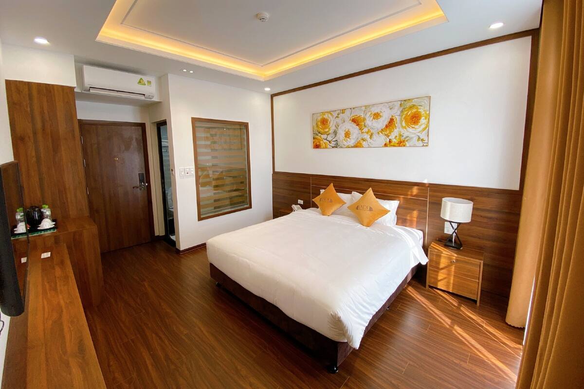 A modern hotel room with a large bed, wooden flooring, a ceiling with recessed lighting, a wall-mounted air conditioner, and a painting above the bed.