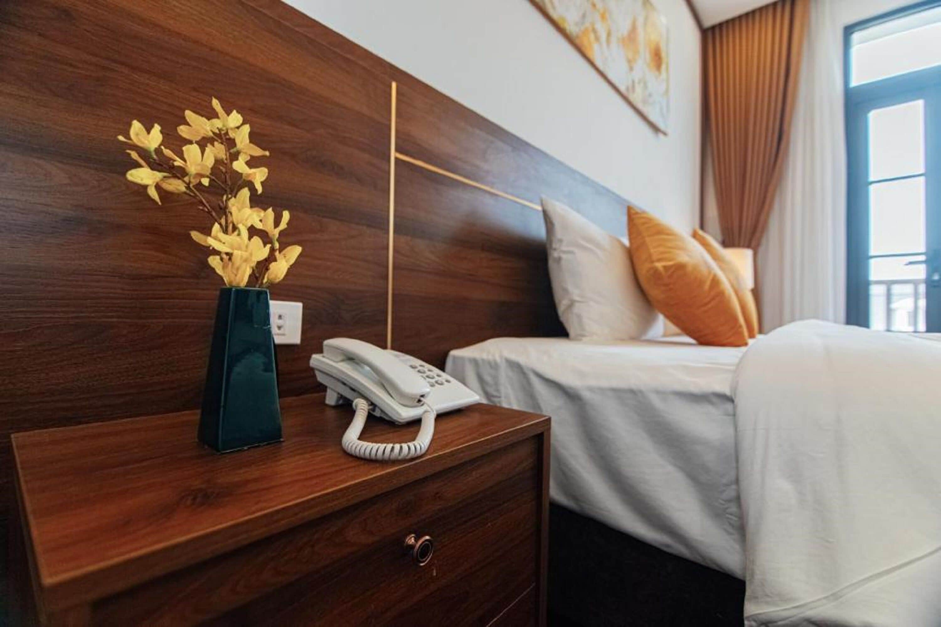 Een hotelkamer met een houten hoofdeinde, een nachtkastje met telefoon, een vaas met bloemen, en een bed met wit beddengoed en oranje kussens.