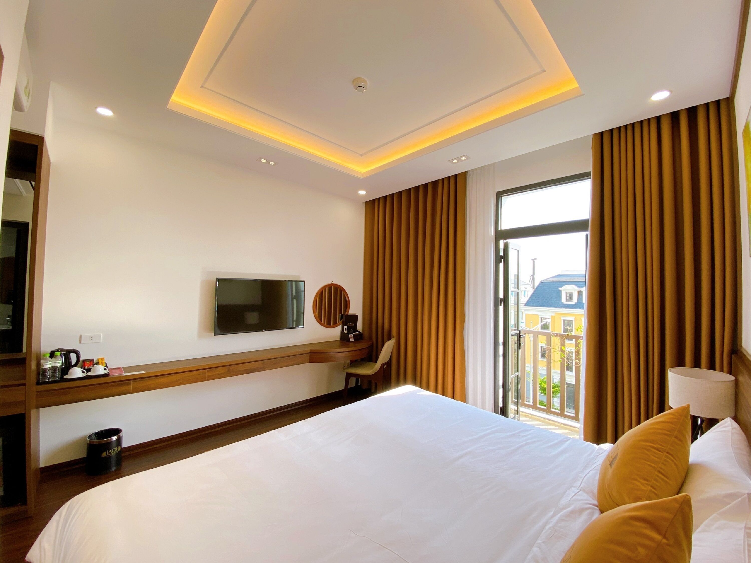 A hotel room with a large bed, a flat-screen TV mounted on the wall, a desk with a chair, and a window with curtains.