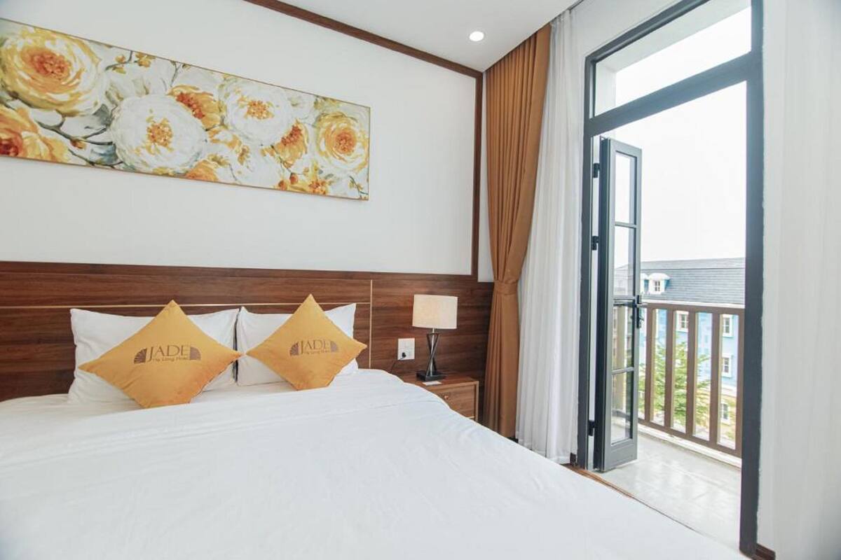 A hotel room with a large bed, two decorative pillows, a nightstand with a lamp, and a balcony with curtains.