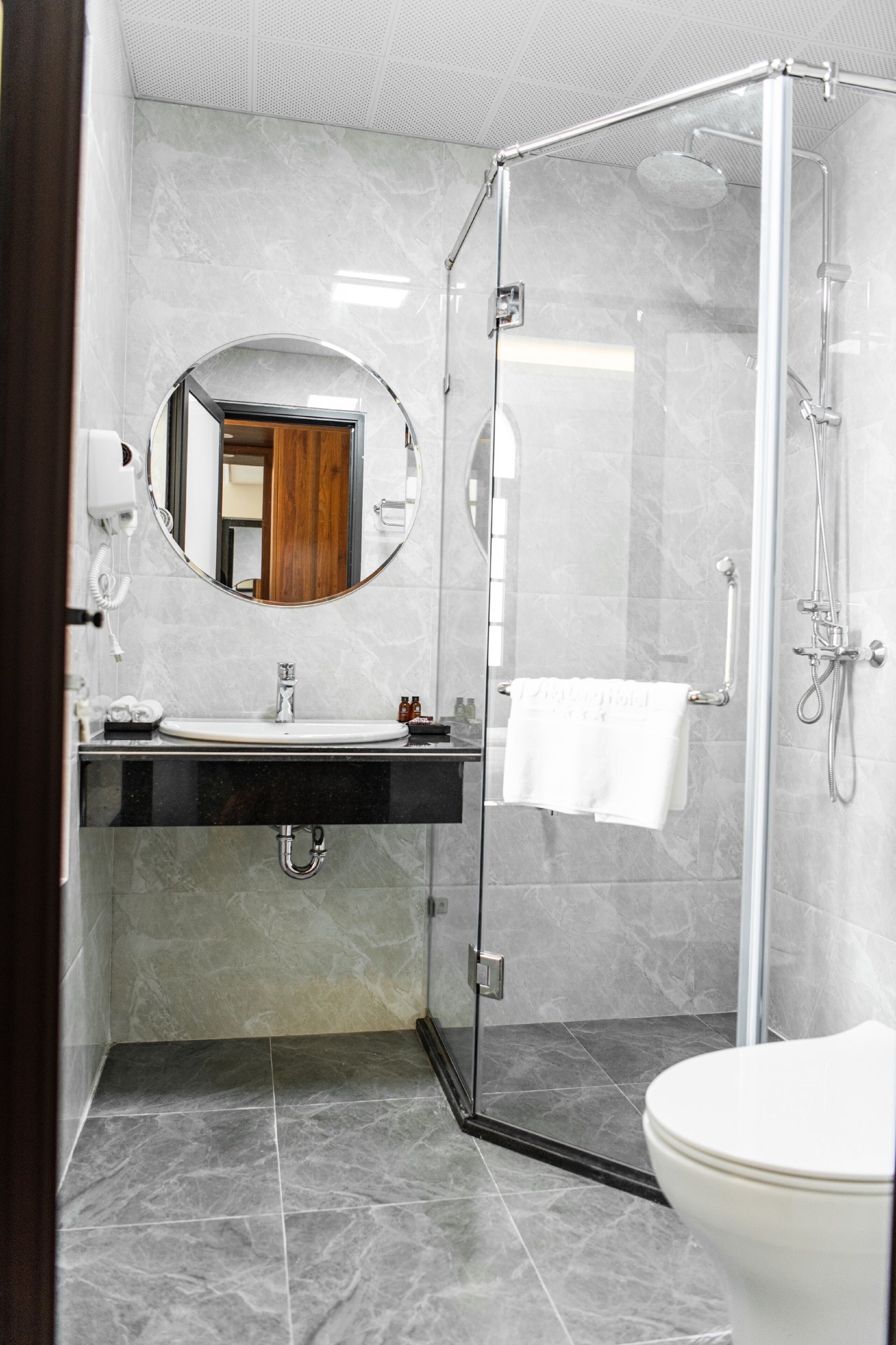 A modern bathroom with a glass-enclosed shower, a round mirror, a sink with a towel rack, and a toilet.