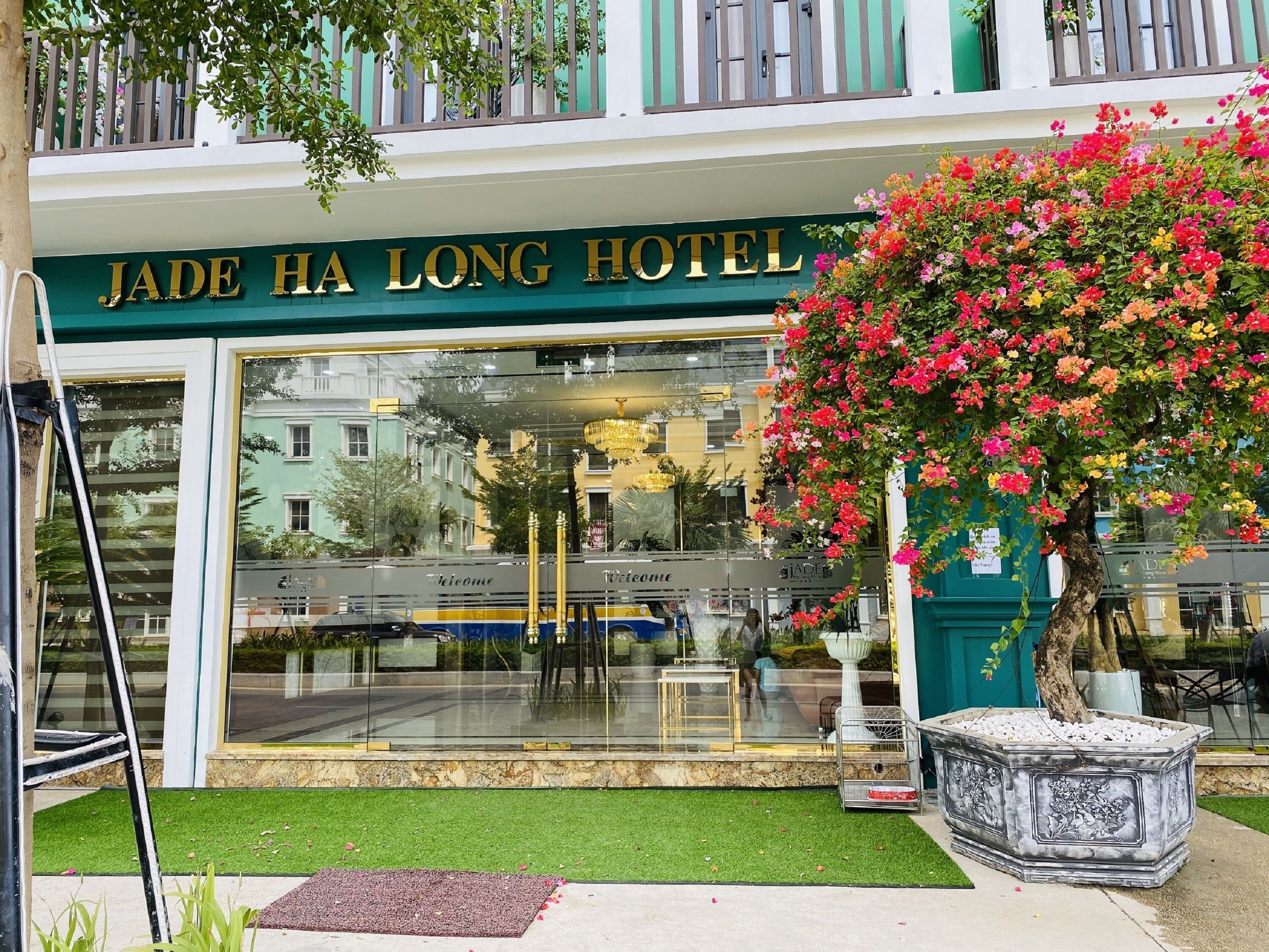 Het uiterlijk van Jade Ha Long Hotel met een grote glazen ingang, een gastvrij bord en een kleurrijke bloemenpresentatie.