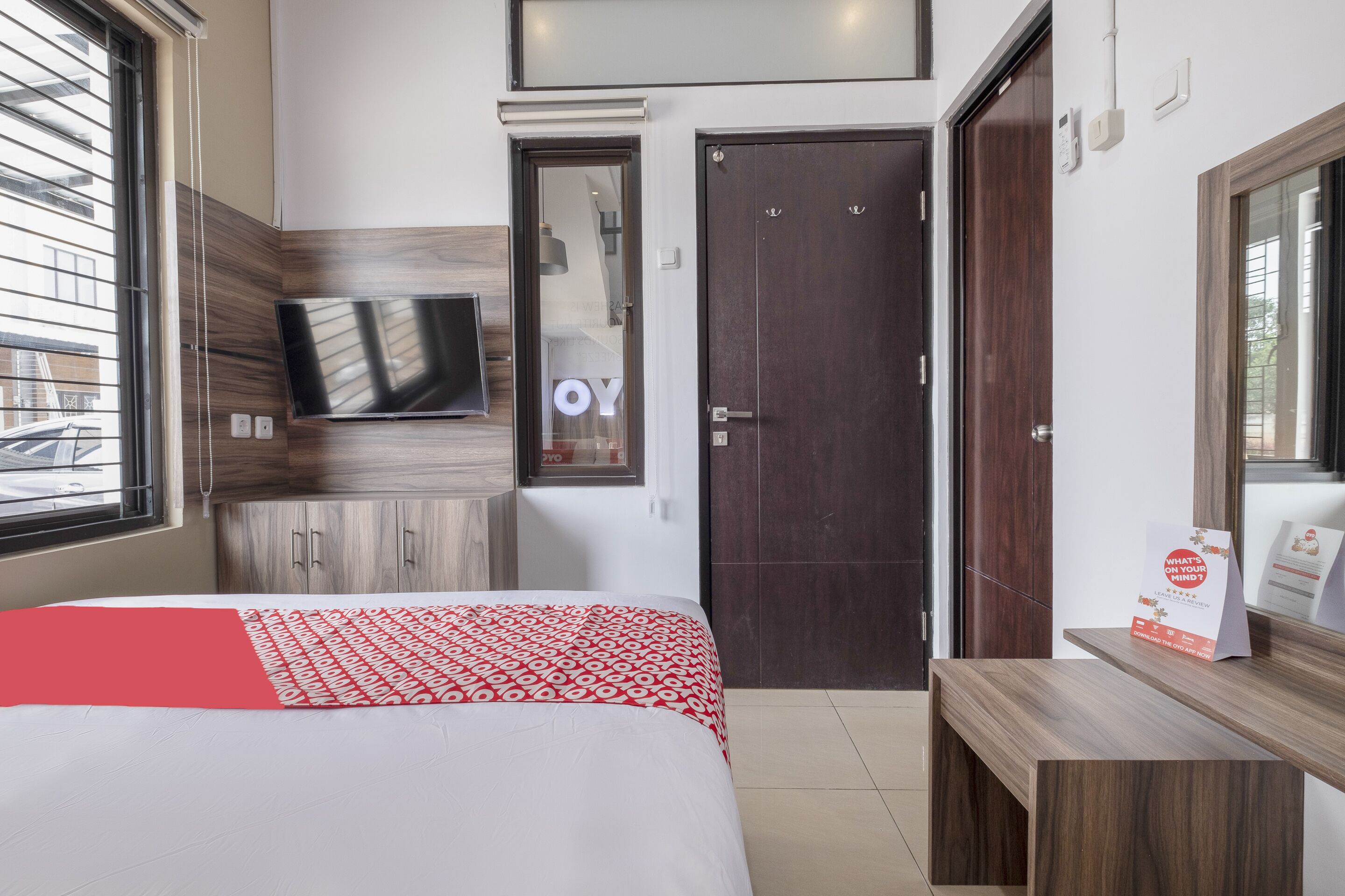 A hotel room with a bed, a TV mounted on the wall, a wooden wardrobe, a dark door, and a bench.