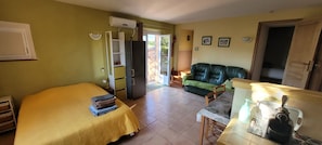 Une pièce comprenant un lit, un canapé vert, une table à manger avec des chaises, un coin cuisine et une porte-fenêtre coulissante donnant sur un espace extérieur.