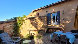 Un bâtiment doté d’une façade en pierre, d’un balcon avec une rambarde en métal et d’un espace extérieur aménagé avec des tables et des chaises.