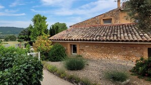 Une maison en pierre avec un toit de tuiles, entourée d’un chemin de gravier et de verdure.