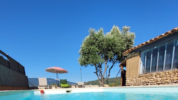 Une piscine d’un bleu éclatant, bordée d’un mur de pierre, avec un arbre et un bâtiment doté d’un toit en tuiles.