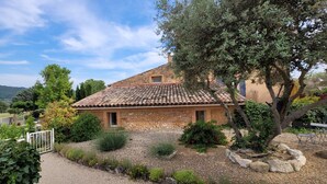 Une maison en pierre avec un toit de tuiles, entourée d’un jardin de gravier et d’arbres.