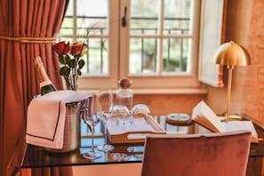 A table set with a bottle of champagne, glasses, a vase with roses, and a book, with a window and curtains in the background. - Château Hôtel - Domaine de Coeurmandie (Bény-sur-Mer)