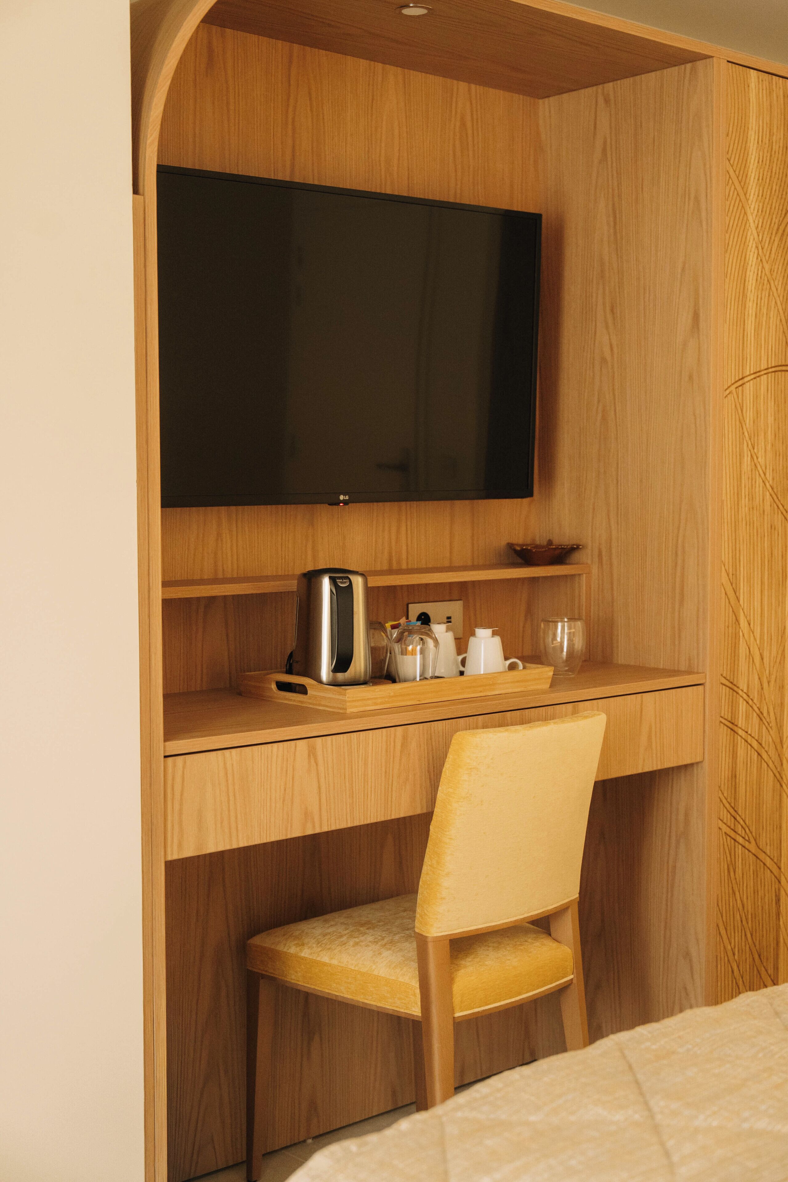 Une pièce de taille modeste, comprenant un bureau en bois, une chaise, un téléviseur à écran plat fixé au mur et une petite étagère avec une bouilloire et des tasses.