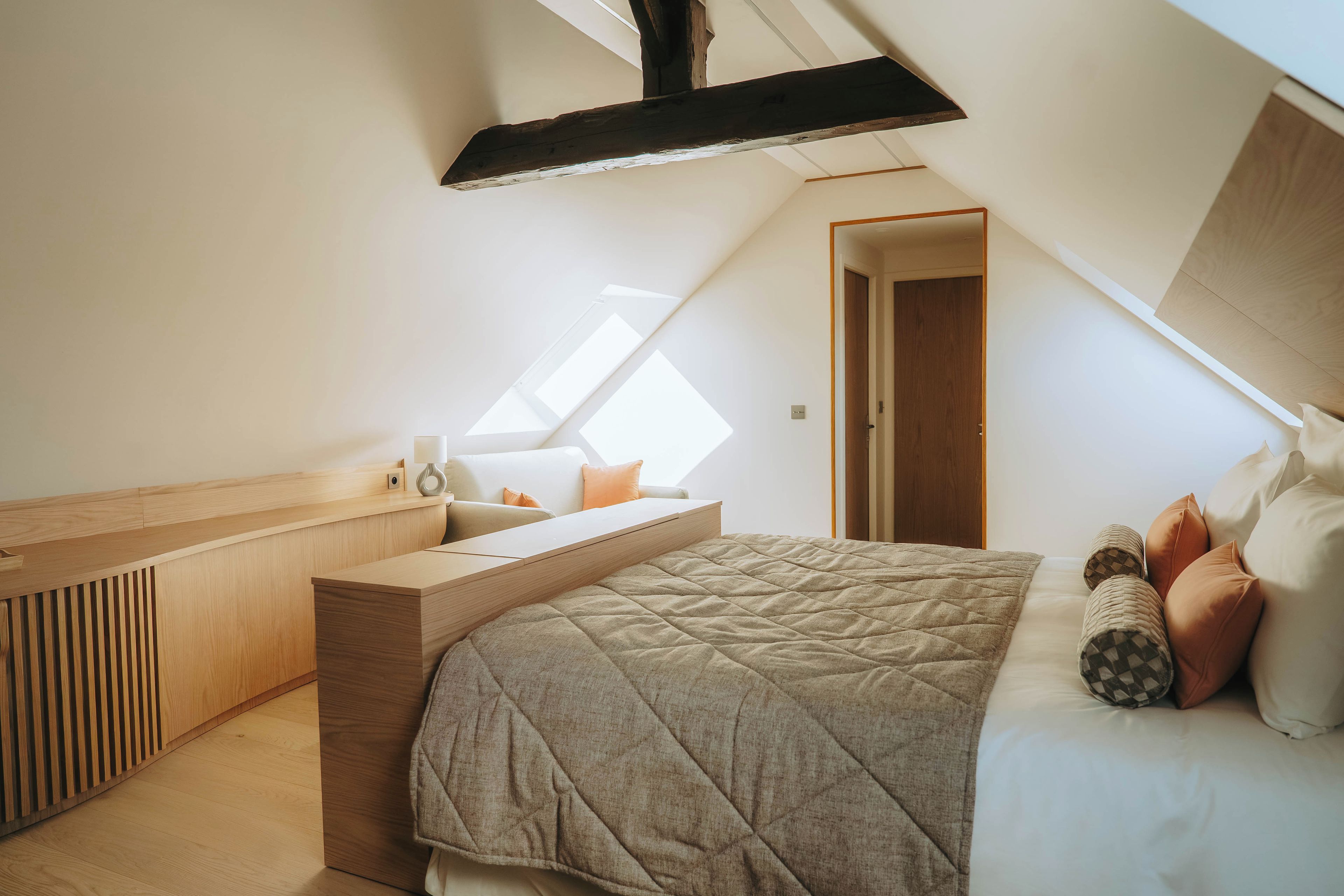 A modern bedroom with a sloped ceiling, a built-in desk, a bed with a quilted cover, and a window providing natural light.