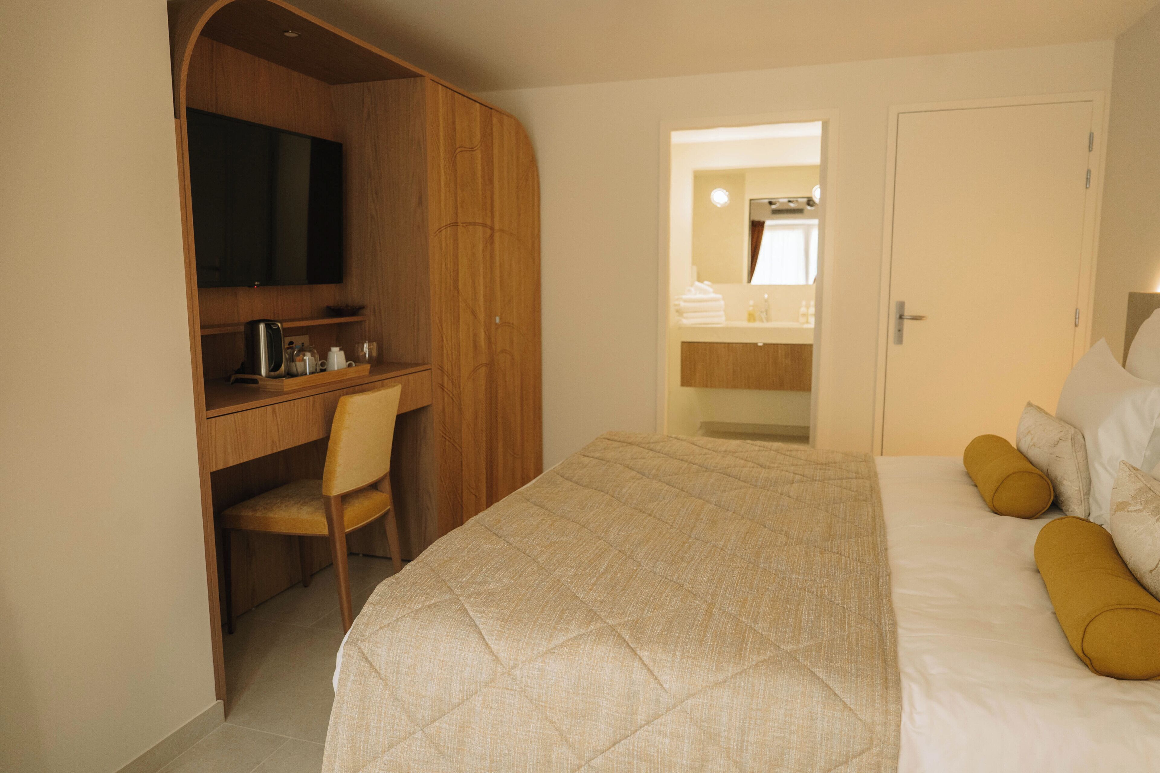 A hotel room with a bed, a desk with a chair, a television, and a bathroom visible through an open door.
