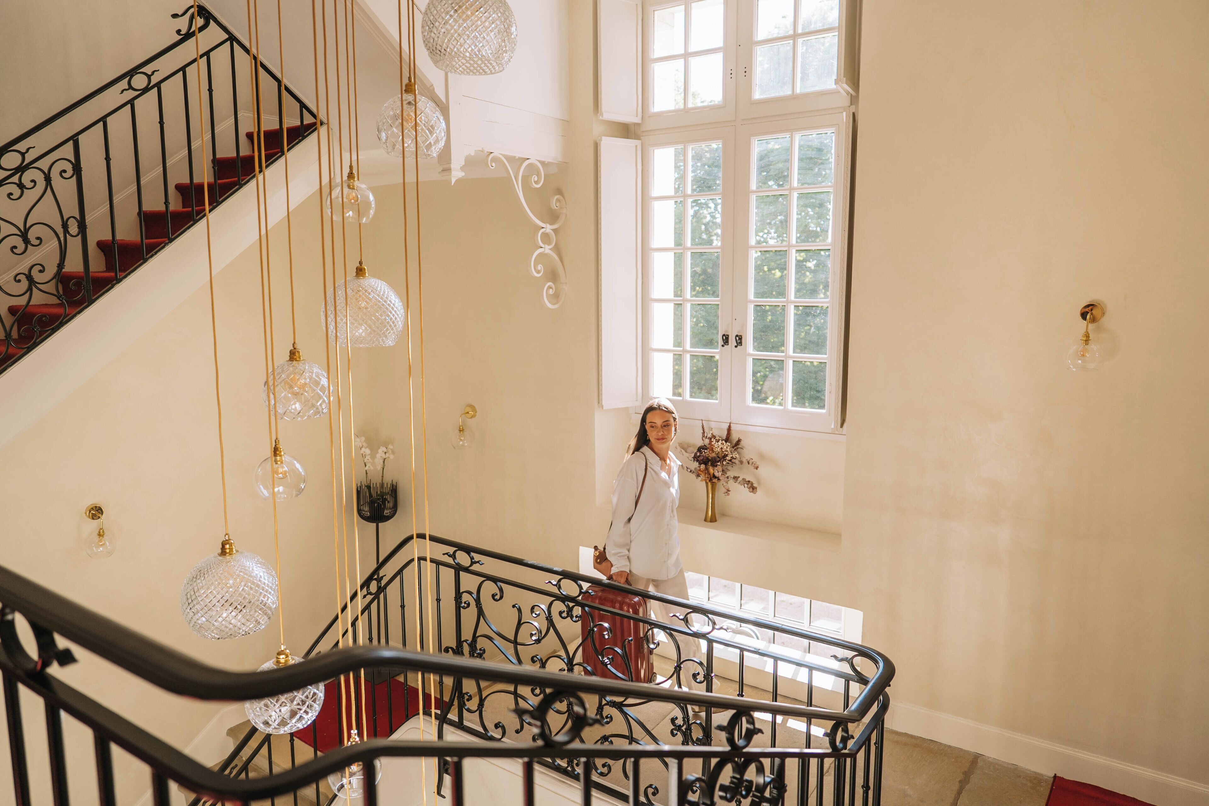 Eine Treppe mit aufwendiger Metallreling und Kristalllüstern, die zu einem Raum mit einem großen Fenster und einer Wandvase mit Blumen führt.