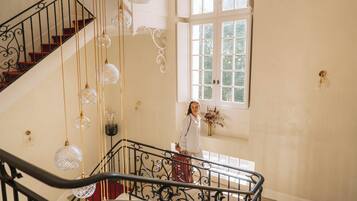 A staircase with ornate metal railings and crystal pendant lights, leading to a room with a large window and a vase of flowers on the wall.
