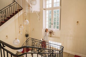 Una escalera con barandillas de metal ornamentadas y lámparas colgantes de cristal, que conduce a un cuarto con un ventanal grande y un florero en la pared.