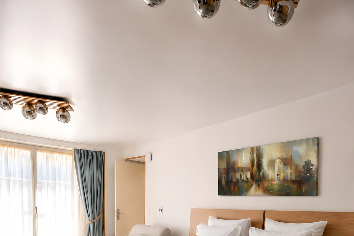 A modern bedroom with a large bed, a painting on the wall, and a view of the outdoors.