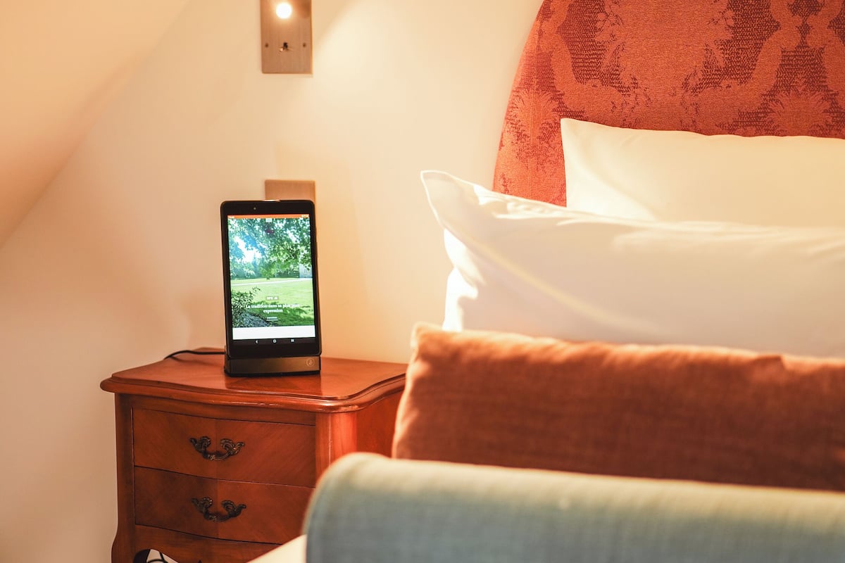 Une chambre à coucher avec un lit, une table de chevet sur laquelle est posée une tablette affichant un paysage, et un luminaire fixé au mur.