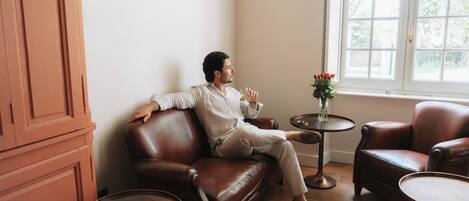 A man seated on a leather sofa in a room with a window, a small table with a vase of flowers, and a cabinet.