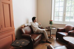 A man seated on a leather sofa in a room with a window, a small table with a vase of flowers, and a cabinet.