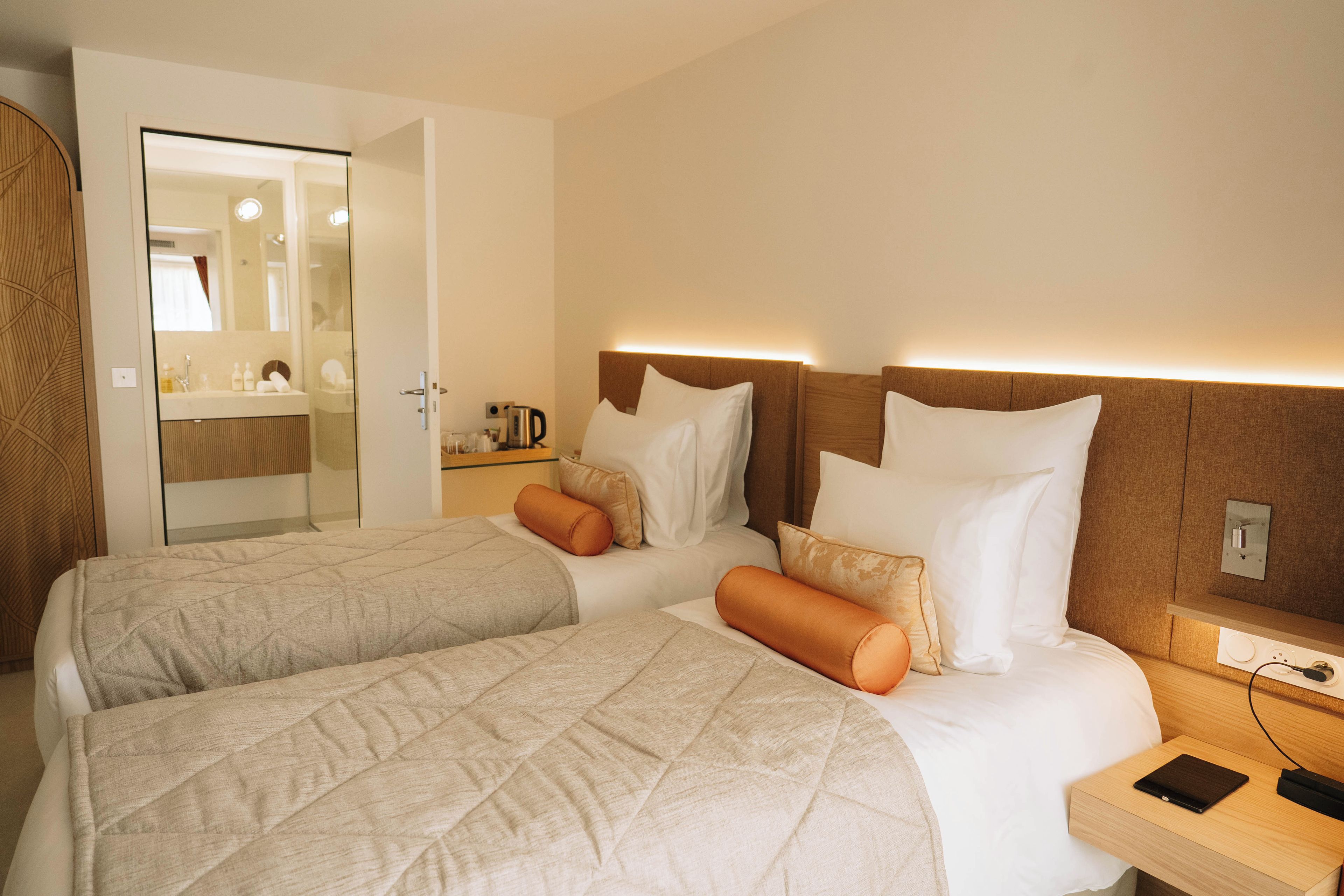A hotel room with two beds, a bedside table with a lamp, a nightstand with a laptop, and a bathroom visible through an open door.