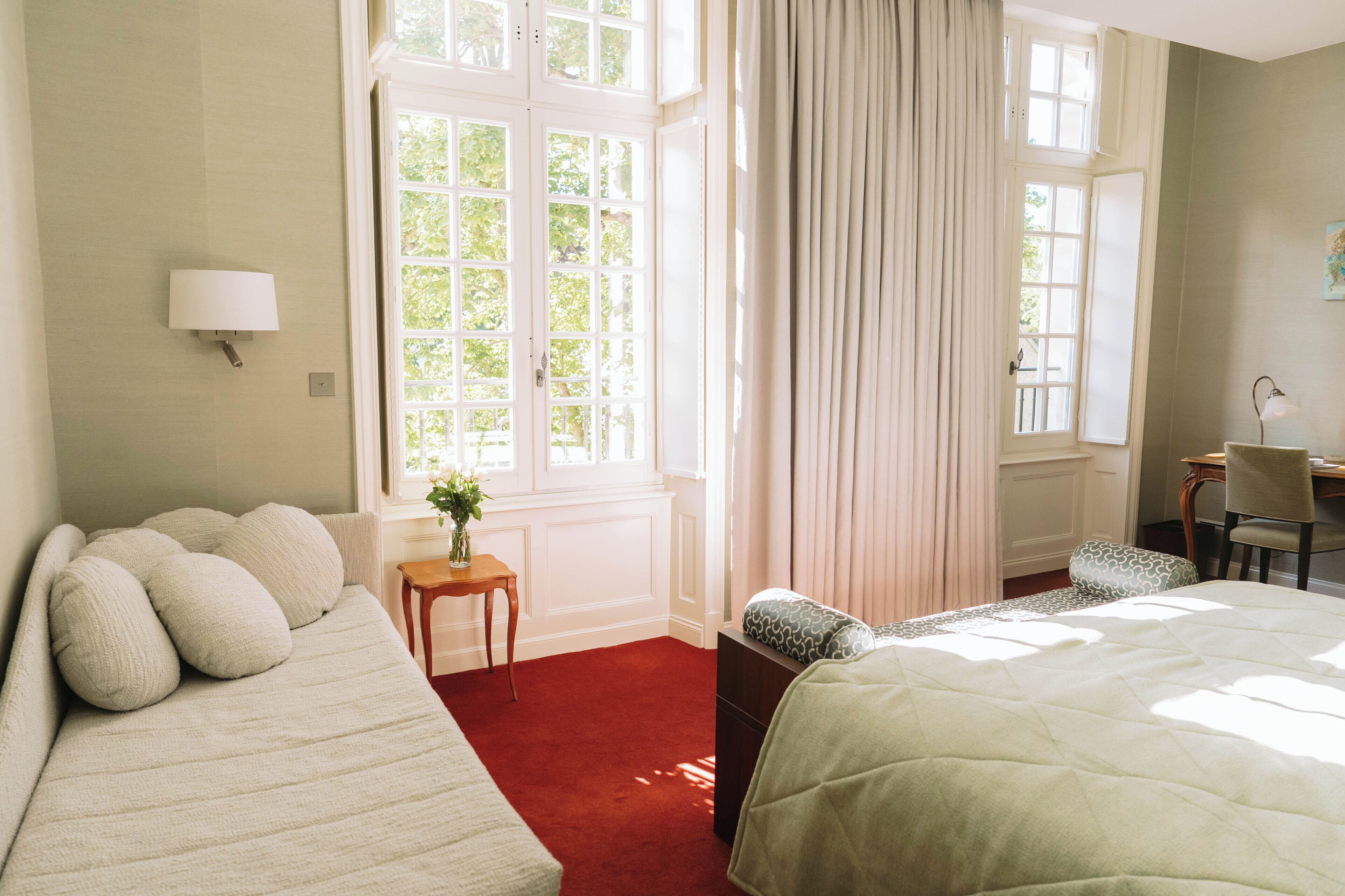 A hotel room with two beds, a desk, a chair, a lamp, and a window with curtains.