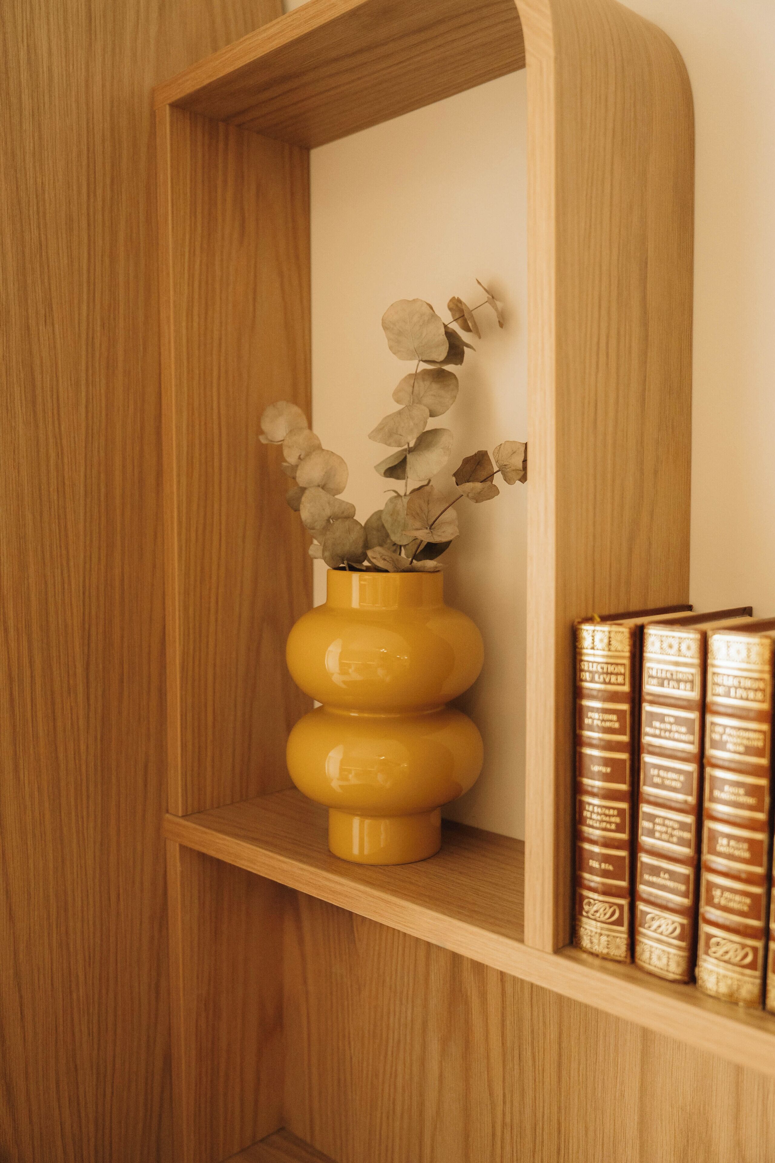 Une étagère en bois sur laquelle se trouve un vase jaune contenant des fleurs séchées, ainsi qu’une rangée de livres.
