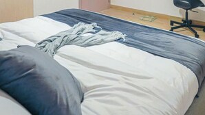 Una cama bien hecha con edredón gris y blanco, una almohada roja y una almohada azul. Junto a la cama, un taburete de madera con una cesta encima.