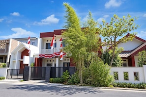Um edifício residencial de dois andares com varanda, uma bandeira e uma árvore em primeiro plano.