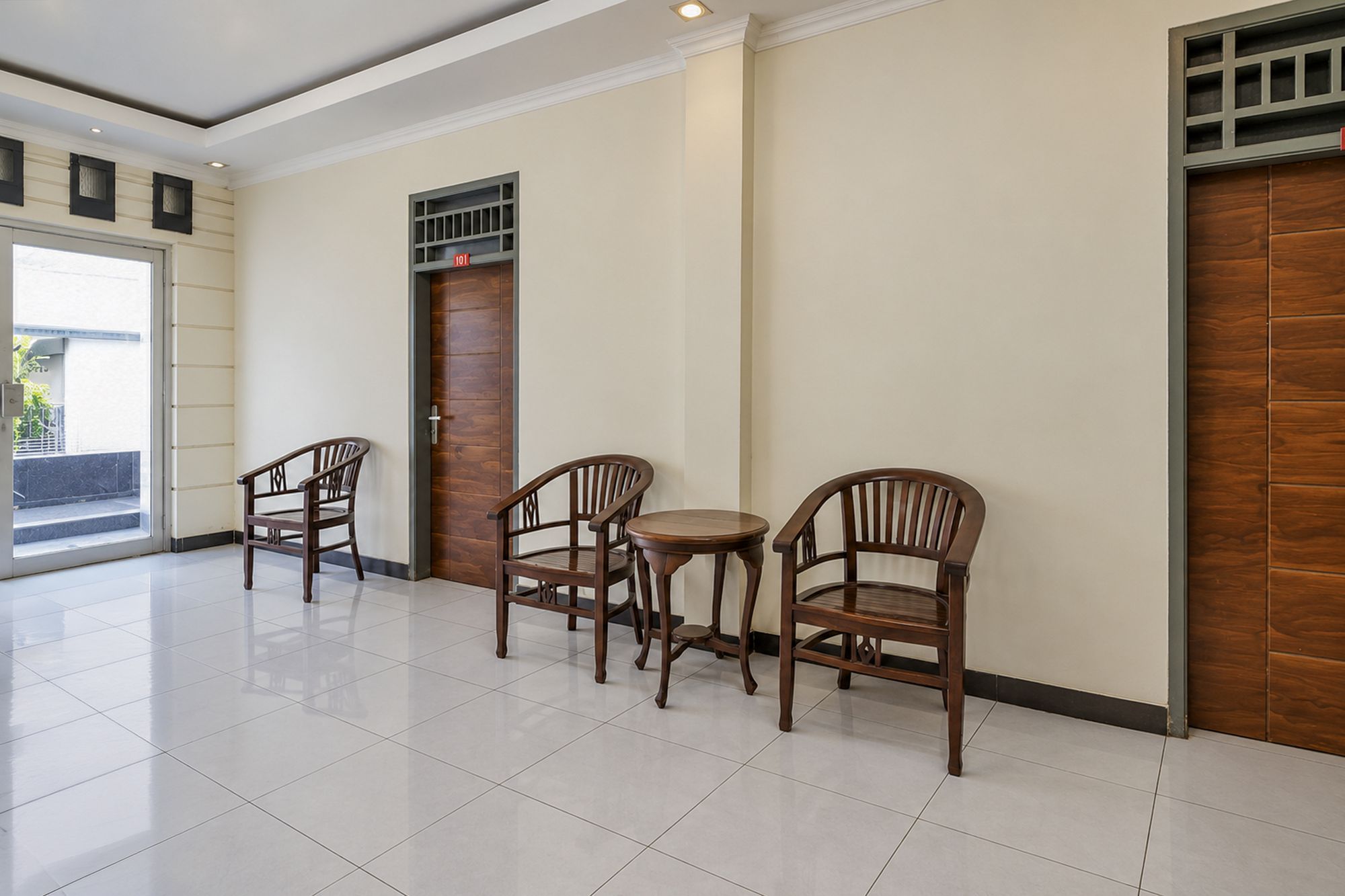 A hotel lobby with a tiled floor, wooden chairs, a small round table, and a door with a security panel.