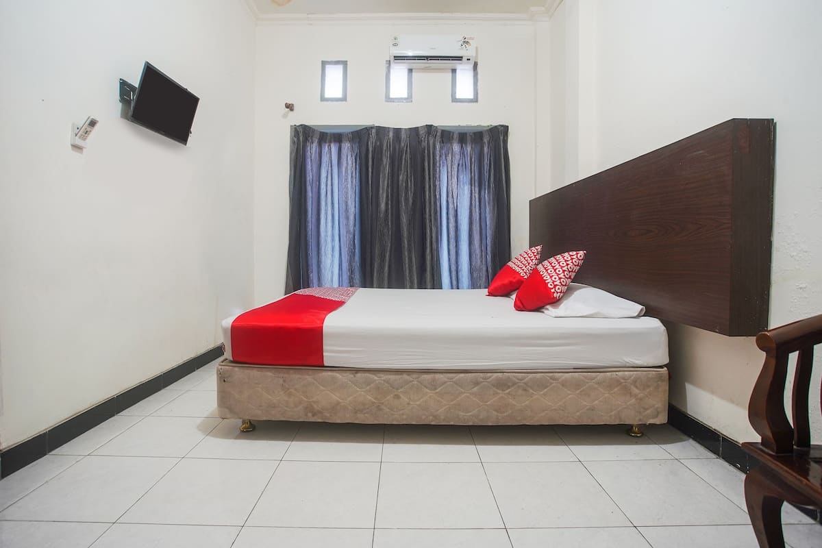 A small, neatly arranged bedroom with a bed, a wooden headboard, a wall-mounted TV, and a window with curtains.