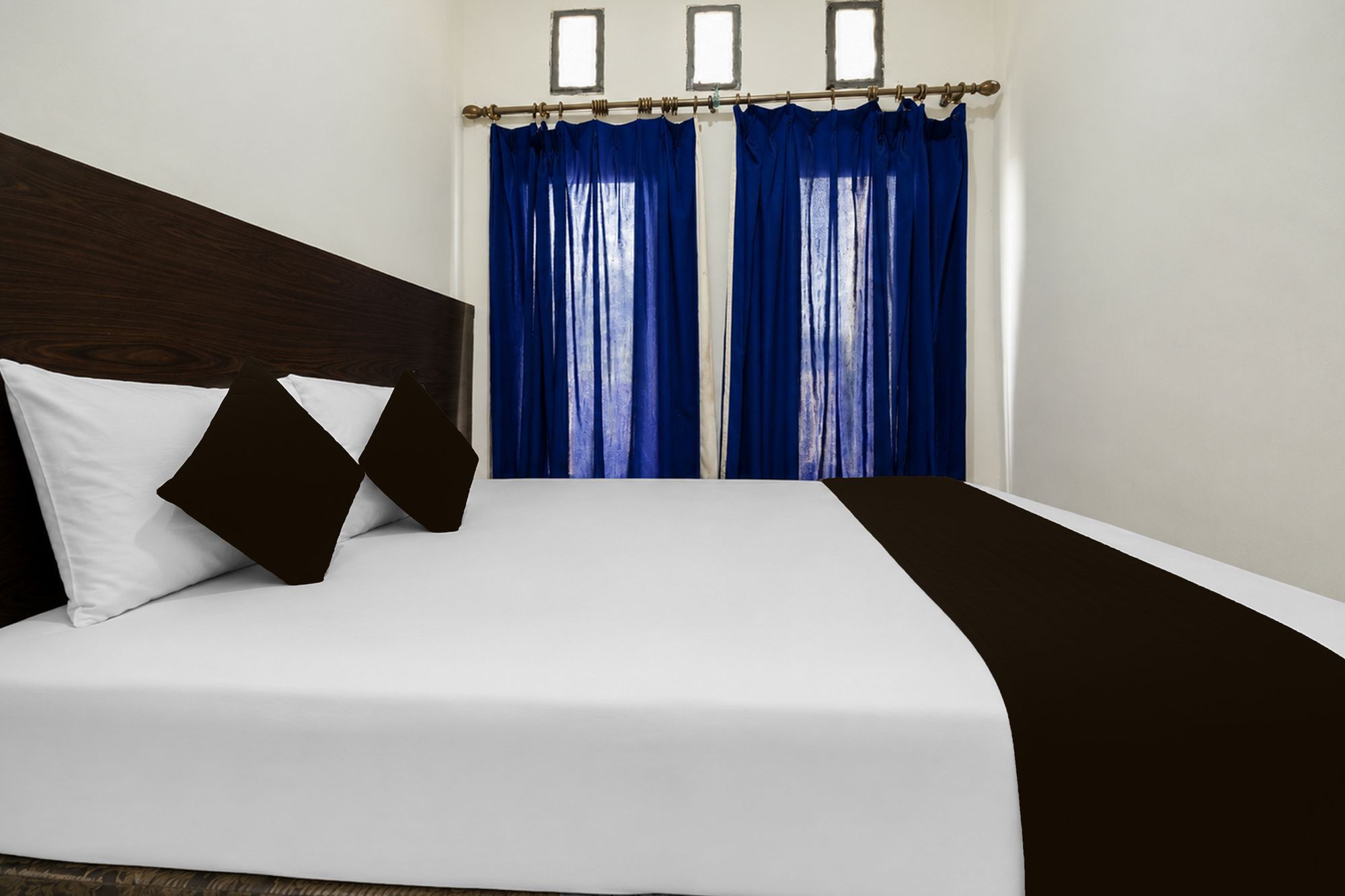 Una habitación de hotel con una cama, ropa de cama blanca, un cabecero oscuro y cortinas azules.